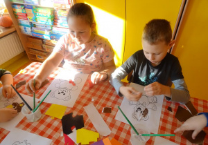 Dwójka dzieci wykleja kolorowymi kawałkami wycinanki szablon misia.